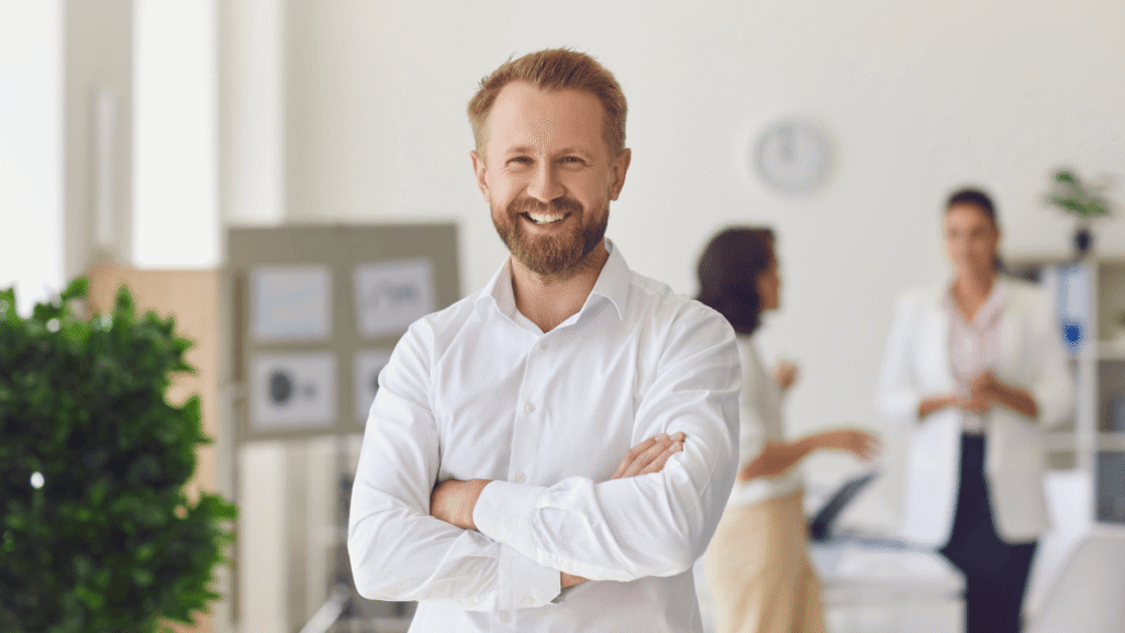 A bearded man in a white shirt stands in an office, looking confidently at the camera.